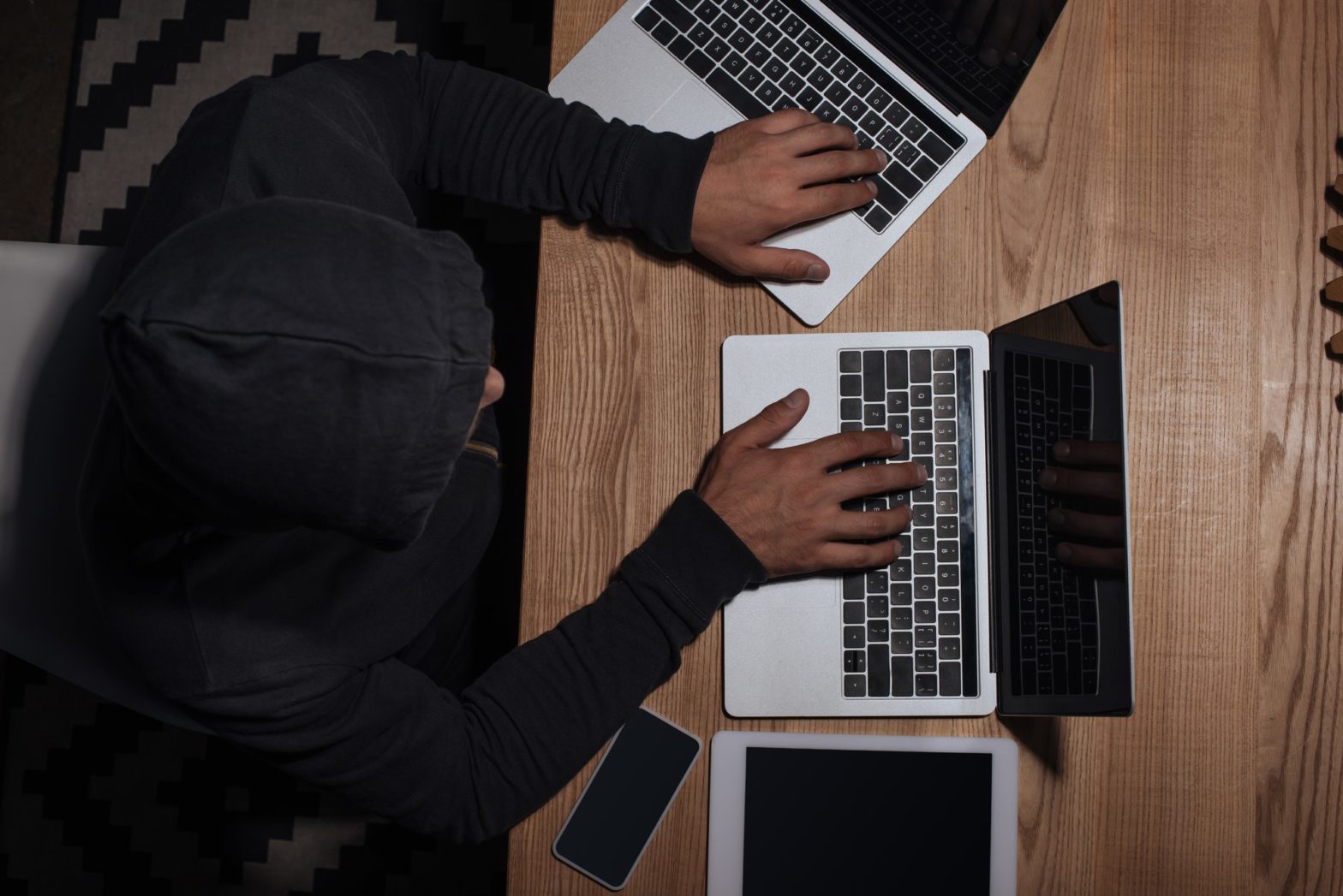 overhead view of hacker in black hoodie using laptops at tabletop with tablet and smartphone, cyber